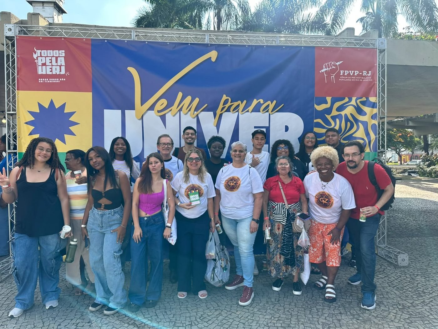Grupo de estudantes e voluntários do IGAZU reunidos em frente a um painel da mobilização 'Vem para a Universidade', em ação pelo acesso à UERJ
